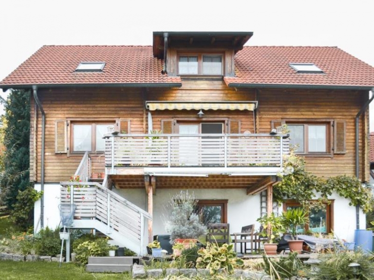 Ein zweistöckiges Holzhaus mit rotem Ziegeldach, das erst kürzlich durch Dachsanierung von einem Experten der Zimmerei aufgefrischt wurde, sowie ein Balkon mit Geländer, eine Außentreppe und ein üppiger Garten mit Topfpflanzen und Terrassenmöbeln vor dem Haus.