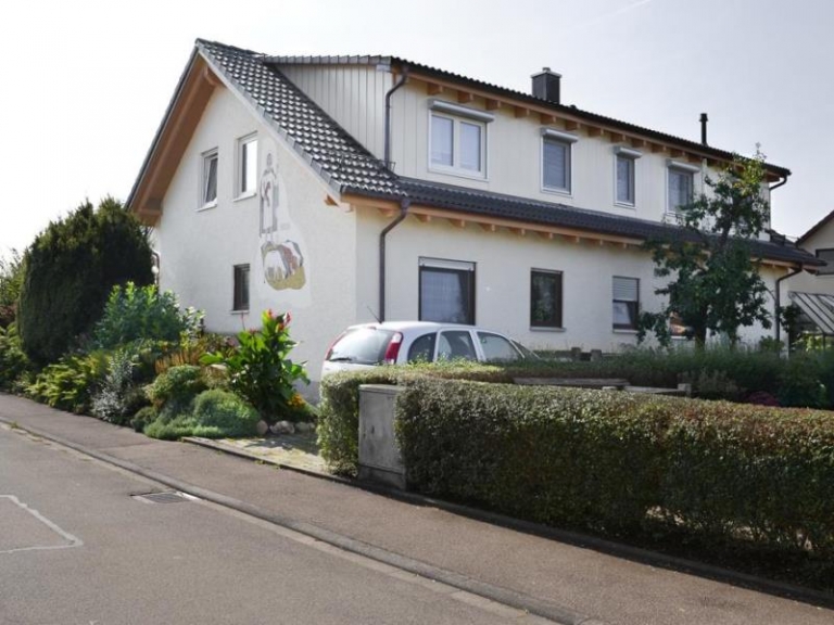 Ein zweistöckiges, cremefarbenes Haus mit einem dunkelgrauen Dach, das durch fachmännische Dachsanierung erneuert wurde, ist von grünen Sträuchern und Pflanzen umgeben. Ein weißes Auto parkt in der Einfahrt, und ein Wandgemälde ziert die Seitenwand entlang einer ruhigen Straße.