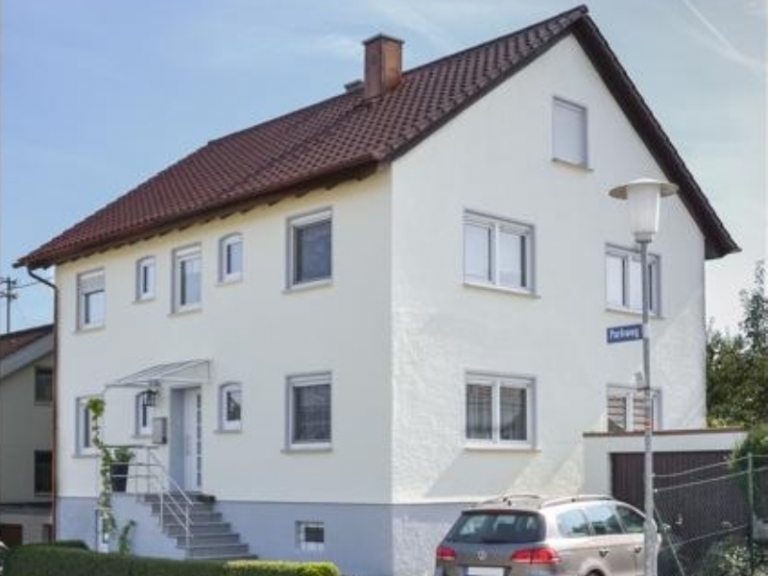 Ein zweistöckiges weißes Haus mit rotem Ziegeldach, das durch fachmännische Dachsanierung renoviert wurde, hat mehrere Fenster und eine kleine Treppe, die zur Eingangstür führt. Davor ist ein silbernes Auto geparkt, und an der Ecke ist ein Straßenschild zu sehen.