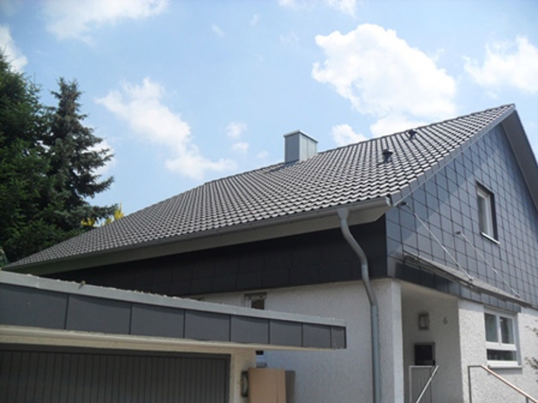 Ein modernes Haus mit steilem, dunklem Ziegeldach und hellen Außenwänden, mit einer Garage im Vordergrund und einem Schornstein auf dem Dach - ideal für Zimmerei-Projekte oder Dachsanierung bei teilweise bewölktem Himmel.