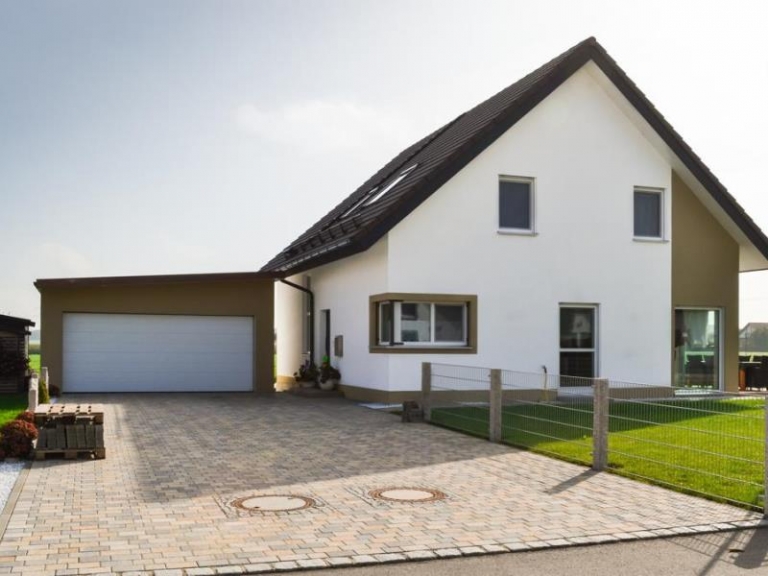 Modernes zweistöckiges weißes Haus mit dunklem Dach, angebauter Garage, gepflasterter Auffahrt, grünem Rasen und Metallzaun an einem sonnigen Tag - fachmännische Zimmerei und Dachsanierung für nachhaltige Qualität.