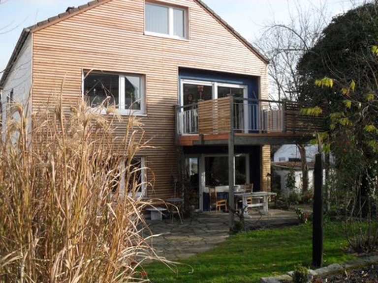 Ein modernes zweistöckiges Haus mit Holzvertäfelung von expert Zimmerei, großen Fenstern, einem oberen Balkon mit Metallgeländer und einer darunter liegenden Terrasse. Das Haus ist von Gras, hohen Pflanzen und Bäumen im Hof umgeben.