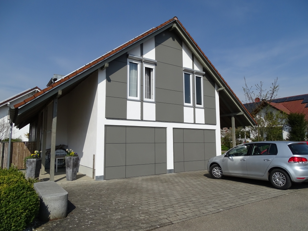Ein modernes Haus mit schrägem Dach, grau-weißer Fassade, großen Garagentoren und zwei hohen Pflanzkübeln neben dem Eingang. Die fachmännische Handwerkskunst der Zimmerei ist überall zu sehen. Ein silbernes Auto parkt auf der gepflasterten Auffahrt unter einem klaren blauen Himmel.