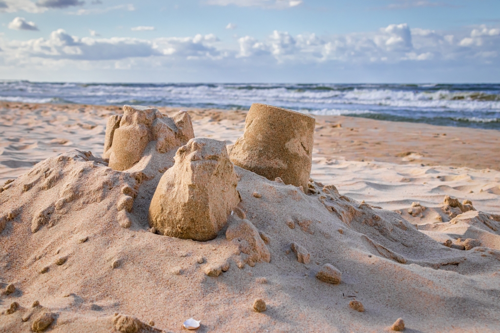 Eine Nahaufnahme von drei teilweise zerbröckelten Sandburgen an einem Sandstrand, die mit der Sorgfalt einer Zimmerei gebaut wurden, mit Wellen und blauem Himmel mit vereinzelten Wolken im Hintergrund.