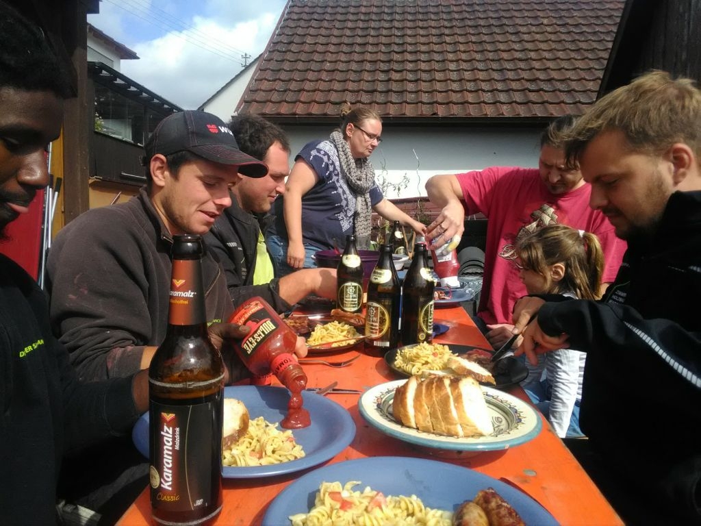 Eine Gruppe von Menschen, darunter auch einige aus einer örtlichen Zimmerei, ist um einen Tisch im Freien versammelt und genießt Bier, Nudeln, Salat und Brot. Einige geben Ketchup auf ihre Teller, während eine Person einem Kind hilft. Die lebhafte Szene spielt sich im Tageslicht ab.