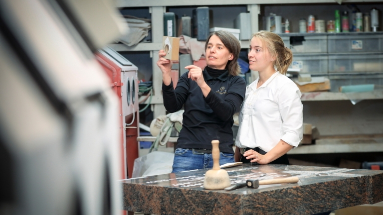 Zwei Frauen stehen in einer Werkstatt. Die eine hält und präsentiert ein kleines Steinstück, während die andere aufmerksam zuhört. Vor ihnen steht ein Granit-Arbeitstisch mit Gravierwerkzeugen, die auf Fertigkeiten hindeuten, die bei Zimmerei- oder sogar Dachsanierungsprojekten nützlich sind.