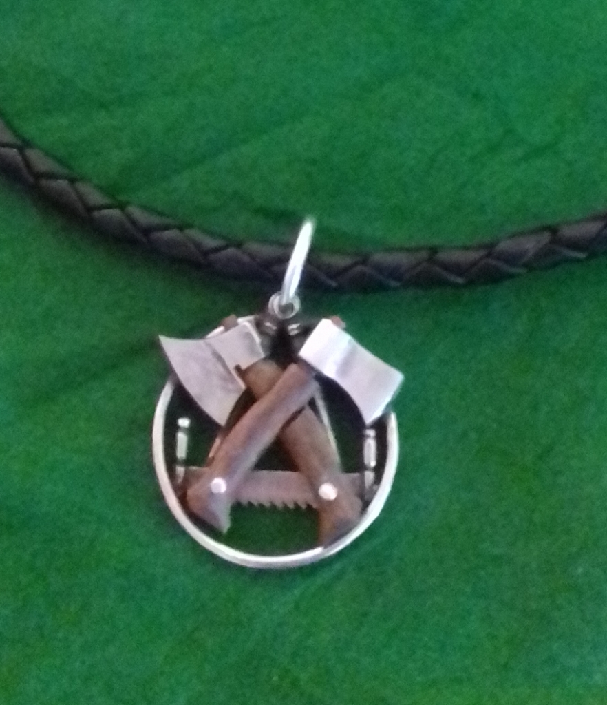 Ein Anhänger mit einer Axt, einer Säge und einem Hammer, die in einem kreisförmigen Rahmen gekreuzt sind - Symbole von Zimmerei -, hängt an einer geflochtenen schwarzen Halskette, die vor einem grünen Hintergrund steht.