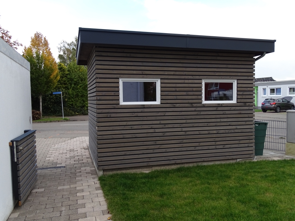 Ein kleiner, moderner Holzschuppen, der mit fachmännischem Zimmerei-Handwerk gebaut wurde, verfügt über zwei rechteckige Fenster neben einer gepflasterten Einfahrt und grünem Rasen, angrenzend an eine Straße und Häuser im Hintergrund.