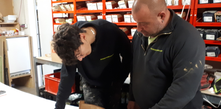 Zwei Personen stehen an einer Werkbank in einer Zimmerei-Werkstatt und beugen sich vor, um etwas zu betrachten, das nicht im Bild ist. Im Hintergrund sind Regale mit roten Behältern und verschiedenen Werkzeugen für die Dachsanierung zu sehen.