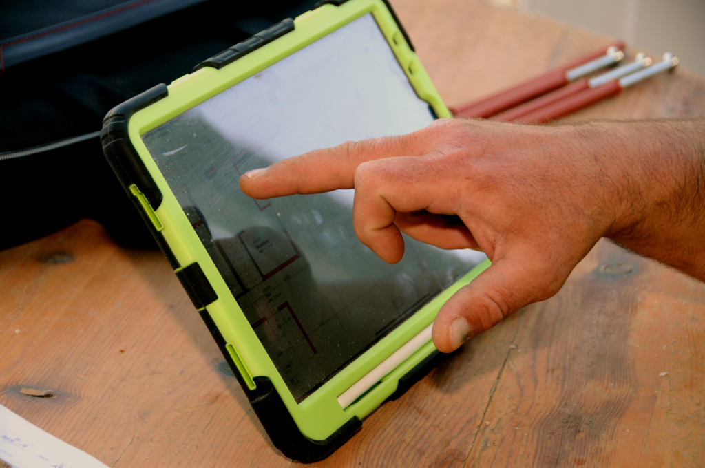 Die Hand einer Person berührt den Bildschirm eines Tablets in einer grünen Schutzhülle, das auf einem Holztisch liegt - wahrscheinlich wird es zur Überprüfung von Zimmerei- oder Dachsanierungsplänen verwendet. Im Hintergrund sind teilweise Metallstangen und eine Tasche zu sehen.