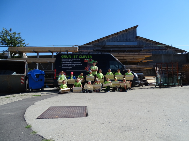 Eine Gruppe von Personen in leuchtend grünen Hemden, wahrscheinlich von einem Zimmerei-Team, sitzt und steht vor einem Lieferwagen und einem Industriegebäude, jeder hält eine Holzkiste. Auf dem Lieferwagen steht GRÜN IST CLEVER. Es ist ein sonniger Tag - perfekt, um über Dachsanierung zu diskutieren.