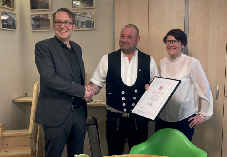 Drei Personen lächeln in einem Haus. Ein Mann in einem dunklen Anzug schüttelt einem anderen, der eine traditionelle Zimmereiweste und ein weißes Hemd trägt, die Hand. Eine Frau mit Brille und weißer Bluse steht daneben und hält eine gerahmte Urkunde für hervorragende Dachsanierung.