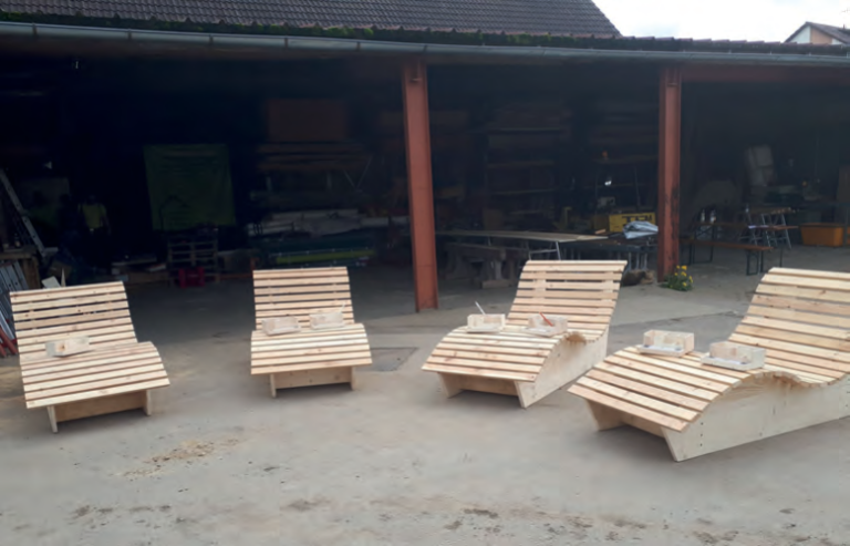 Vier hölzerne Loungesessel mit Lattenrost sind im Freien vor einer Zimmerei-Werkstatt aufgestellt. Jeder Stuhl hat einen kleinen Holzklotz auf der Sitzfläche, und im Hintergrund sind Holzbearbeitungsgeräte für die Dachsanierung zu sehen.