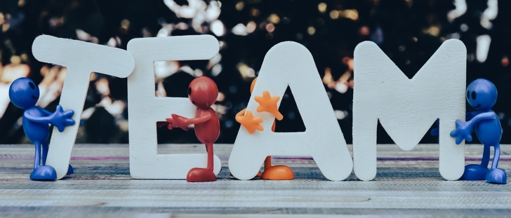 Vier bunte Plastikfiguren halten große weiße Buchstaben hoch, die TEAM auf einer hölzernen Oberfläche buchstabieren und Teamwork und Zusammenarbeit vor einem verschwommenen Hintergrund symbolisieren.