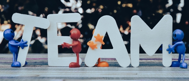 Vier bunte Plastikfiguren halten große weiße Buchstaben hoch, die TEAM auf einer hölzernen Oberfläche buchstabieren und Teamwork und Zusammenarbeit vor einem verschwommenen Hintergrund symbolisieren.