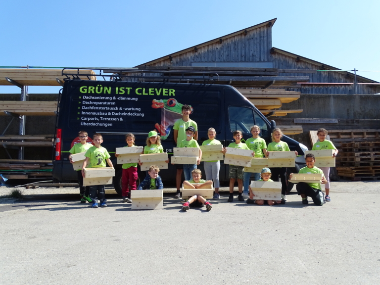 Eine Gruppe von Kindern und zwei Erwachsenen, die alle die gleichen grünen Hemden tragen, stehen im Freien vor einem schwarzen Lieferwagen und einem Holzgebäude, jeder hält eine Holzkiste und lächelt - sie feiern die Teamarbeit bei einer Veranstaltung der Zimmerei Dachsanierung.
