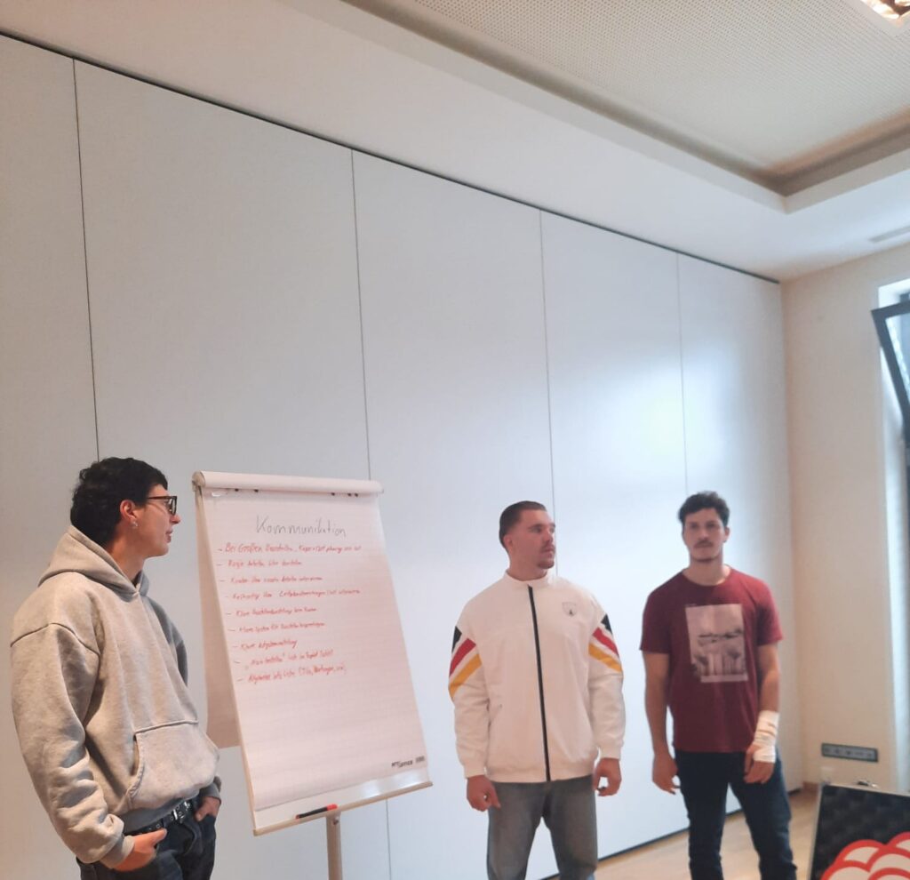Drei Personen stehen in einem Raum neben einem Flipchart mit handschriftlichen Notizen über die Zimmerei. Einer trägt einen grauen Kapuzenpulli, ein anderer eine weiße Jacke mit Streifen und der dritte ein rotes T-Shirt. Der Raum hat weiße Wände und ein großes Fenster.