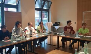 Sechs Personen sitzen um einen Konferenztisch in einem hellen Besprechungsraum und hören einem Zimmereiexperten aufmerksam zu. Auf dem Tisch stehen Wasserflaschen, Gläser und Notizblöcke. Große Fenster lassen natürliches Licht herein.