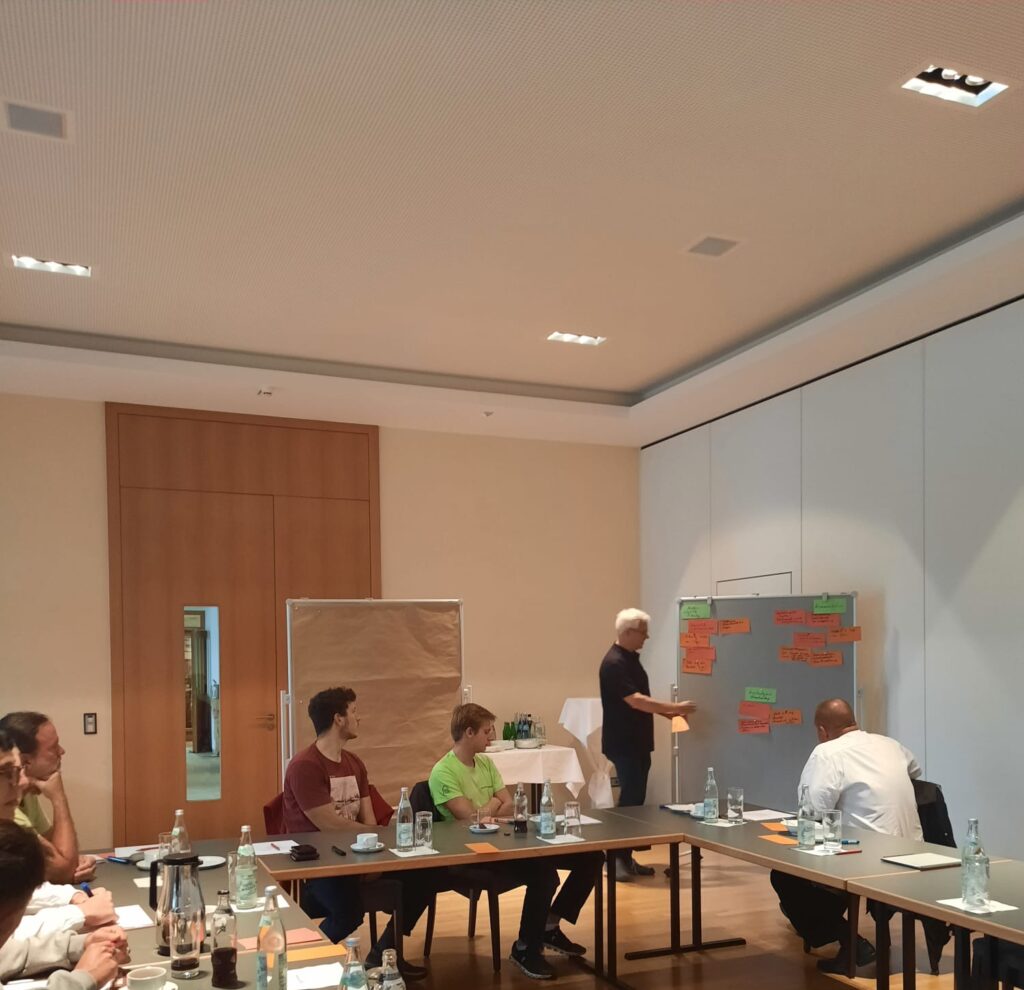 Eine Gruppe von Menschen sitzt in einem Konferenzraum um Tische herum und beobachtet einen Mann, der an einer Tafel mit bunten Haftnotizen arbeitet. Flaschen und Papiere stehen auf den Tischen. Der Workshop scheint sich auf Themen wie Dachsanierung oder Zimmerei zu konzentrieren.